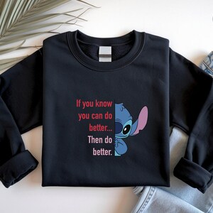 May include: Black sweatshirt featuring a blue and pink cartoon character and the text "If you know you can do better... Then do better." The design is on the front of the long-sleeved crew neck top. A palm frond and denim jeans are in the background.