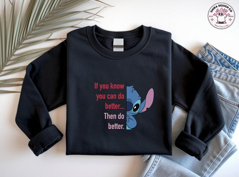May include: Black sweatshirt featuring a blue and pink cartoon character and the text "If you know you can do better... Then do better." The design is on the front of the long-sleeved crew neck top. A palm frond and denim jeans are in the background.