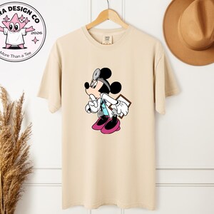 May include: Beige t-shirt featuring a cartoon Mickey Mouse dressed as a doctor, wearing a stethoscope and holding a clipboard. The shirt has a round neck and short sleeves. The design includes black, white, pink, and blue colors. The shirt is hanging on a wooden hanger.