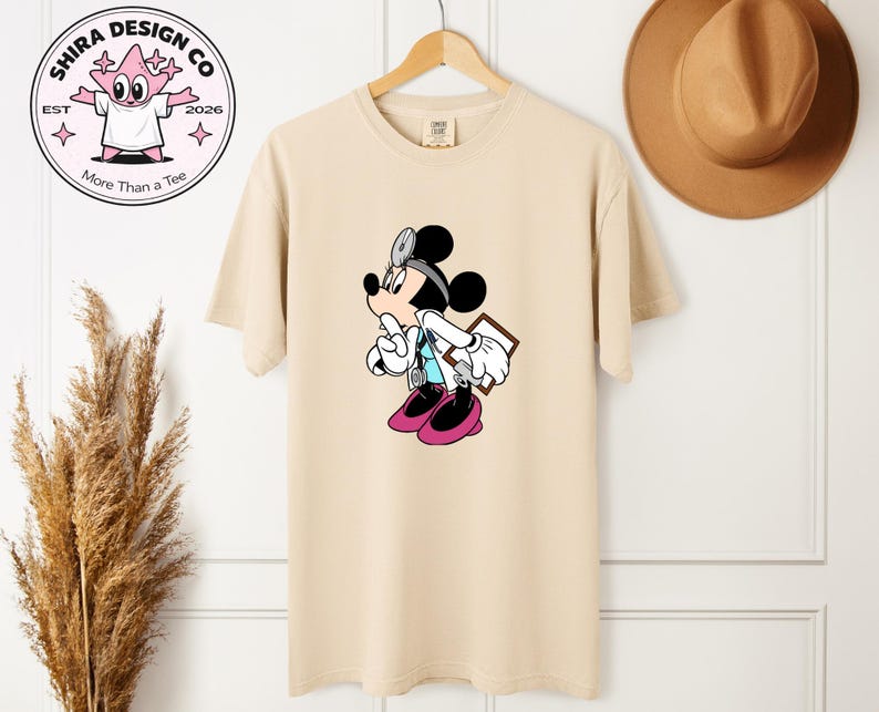 May include: Beige t-shirt featuring a cartoon Mickey Mouse dressed as a doctor, wearing a stethoscope and holding a clipboard. The shirt has a round neck and short sleeves. The design includes black, white, pink, and blue colors. The shirt is hanging on a wooden hanger.