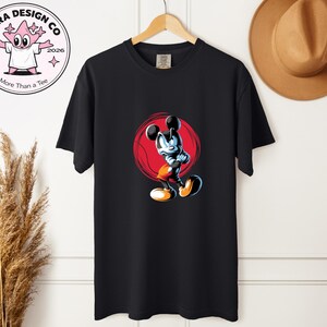 May include: Black t-shirt with a cartoon Mickey Mouse design inside a red circle. The shirt is on a wooden hanger. A brown hat hangs on the wall. The shirt has the text "Shira Design Co" and "More Than a Tee".