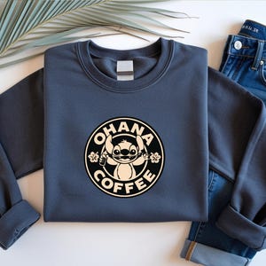 May include: A navy blue sweatshirt featuring a circular graphic with the words "OHANA COFFEE" around a cartoon character. The character is holding a coffee cup and surrounded by flowers. The sweatshirt is paired with blue jeans.