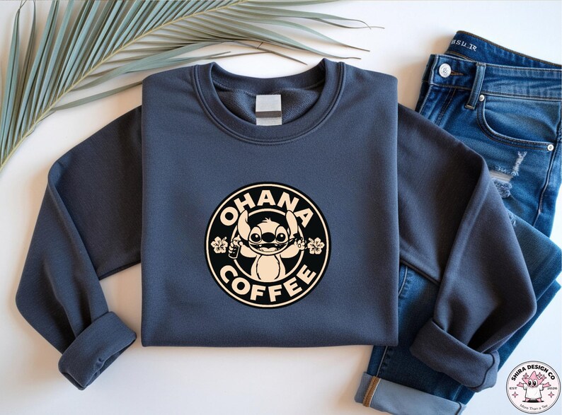May include: A navy blue sweatshirt featuring a circular graphic with the words "OHANA COFFEE" around a cartoon character. The character is holding a coffee cup and surrounded by flowers. The sweatshirt is paired with blue jeans.