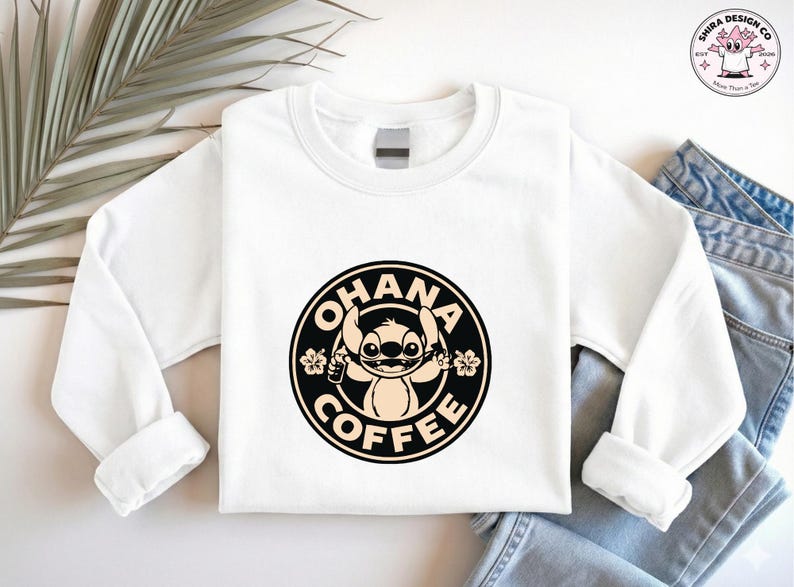 May include: White sweatshirt featuring a black and tan graphic of Stitch from Lilo & Stitch, with the words "OHANA COFFEE" in a circular design. The sweatshirt has long sleeves and a crew neck. The design includes floral accents.