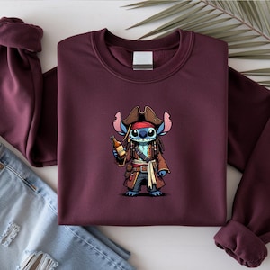 May include: Burgundy crewneck sweatshirt featuring a cartoon pirate character holding a bottle. The character is blue with a red bandana, brown hat, and a brown coat. The sweatshirt is folded with a pair of blue jeans.