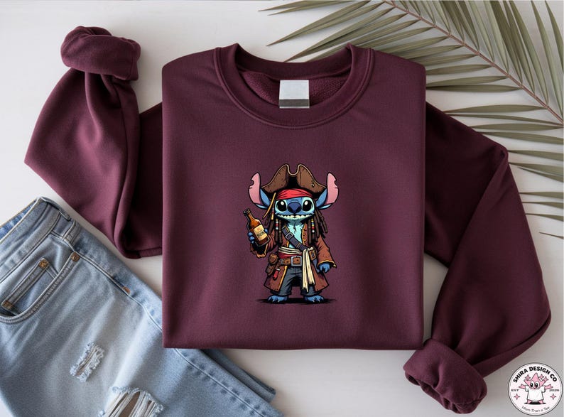 May include: Burgundy crewneck sweatshirt featuring a cartoon pirate character holding a bottle. The character is blue with a red bandana, brown hat, and a brown coat. The sweatshirt is folded with a pair of blue jeans.