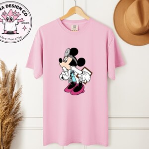 May include: A light pink t-shirt featuring a cartoon Mickey Mouse dressed as a doctor, holding a clipboard and stethoscope. The shirt has the text "Shira Design Co" and "More Than a Tee" on a circular logo.