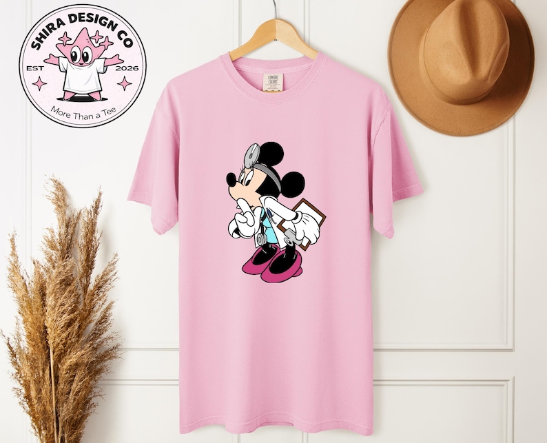 May include: A light pink t-shirt featuring a cartoon Mickey Mouse dressed as a doctor, holding a clipboard and stethoscope. The shirt has the text "Shira Design Co" and "More Than a Tee" on a circular logo.