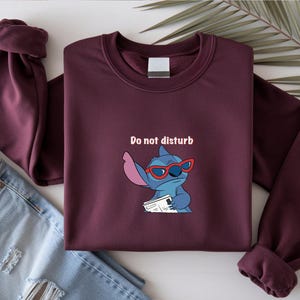 Peut inclure: Sweat-shirt bordeaux avec un personnage de dessin anim&eacute; Stitch portant des lunettes rouges et tenant un papier, avec le texte "Do not disturb". Le sweat-shirt est pli&eacute; avec un jean bleu.