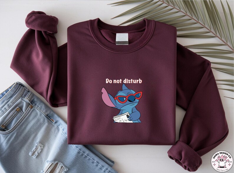 Peut inclure: Sweat-shirt bordeaux avec un personnage de dessin anim&eacute; Stitch portant des lunettes rouges et tenant un papier, avec le texte "Do not disturb". Le sweat-shirt est pli&eacute; avec un jean bleu.