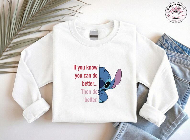 May include: White long-sleeve sweatshirt featuring a blue cartoon character and the text "If you know you can do better... Then do better." The design includes a logo in the upper right corner. The sweatshirt is paired with blue jeans.