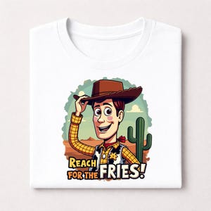 Op de afbeelding: Wit T-shirt met een cartoon cowboy illustratie. De cowboy draagt een bruine hoed, geel geruit overhemd en rode halsdoek. De tekst "REACH FOR THE FRIES!" wordt weergegeven tegen een woestijnachtergrond met een cactus.