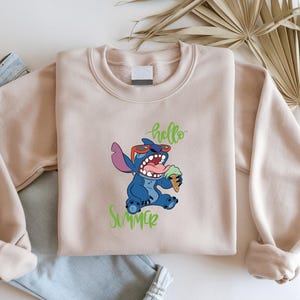 May include: A beige sweatshirt with a blue cartoon character wearing sunglasses and holding an ice cream cone. The words "hello" and "summer" are written in green script. The sweatshirt is folded on top of a pair of blue jeans.