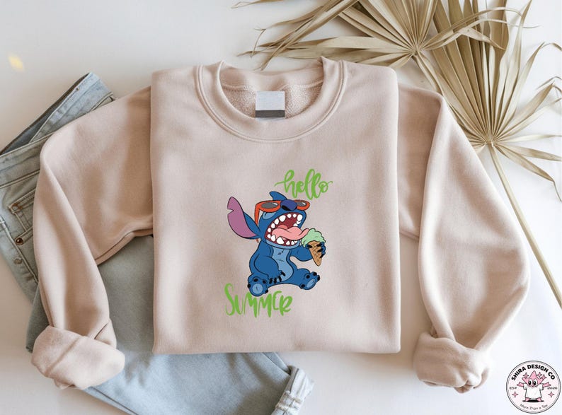 May include: A beige sweatshirt with a blue cartoon character wearing sunglasses and holding an ice cream cone. The words "hello" and "summer" are written in green script. The sweatshirt is folded on top of a pair of blue jeans.
