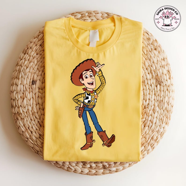 May include: A yellow t-shirt featuring a cartoon illustration of Woody from Toy Story. Woody is wearing a brown cowboy hat, a yellow plaid shirt, blue jeans, and brown boots. The shirt has the text "Shira Design Co" on the upper right corner.
