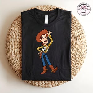 May include: Black t-shirt featuring a cartoon illustration of Woody from Toy Story. Woody is depicted in his signature cowboy attire, including a brown hat, yellow plaid shirt, blue jeans, and brown boots. The shirt also has the text "Shira Design Co" and a cartoon character.