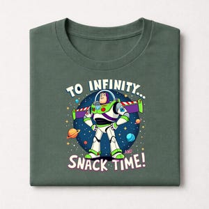 Peut inclure: T-shirt pli&eacute; vert fonc&eacute; avec un motif de Buzz l'&Eacute;clair et le texte "TO INFINITY... AND SNACK TIME!". Le motif repr&eacute;sente une sc&egrave;ne spatiale avec des plan&egrave;tes et des &eacute;toiles, et Buzz est dans sa pose embl&eacute;matique.