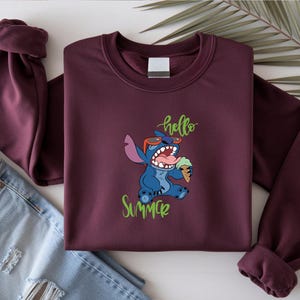 May include: A maroon sweatshirt with a graphic of Stitch from Lilo & Stitch, wearing sunglasses and holding ice cream. The words "hello" and "Summer" are in green script. A pair of blue jeans is visible.