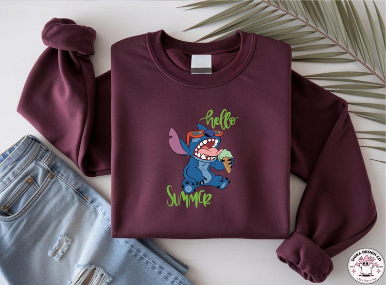May include: A maroon sweatshirt with a graphic of Stitch from Lilo & Stitch, wearing sunglasses and holding ice cream. The words "hello" and "Summer" are in green script. A pair of blue jeans is visible.