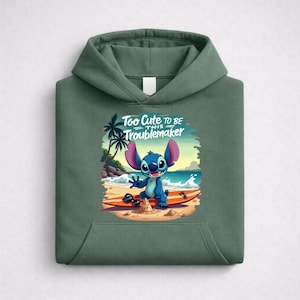 Op de afbeelding: Groene hoodie met een afbeelding van Stitch uit Lilo & Stitch op een surfplank op het strand. De afbeelding bevat de tekst "Too Cute to be this Troublemaker".