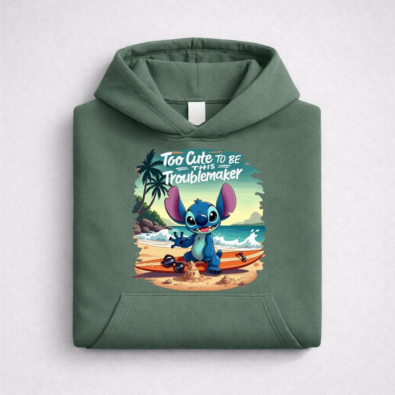 Op de afbeelding: Groene hoodie met een afbeelding van Stitch uit Lilo & Stitch op een surfplank op het strand. De afbeelding bevat de tekst "Too Cute to be this Troublemaker".