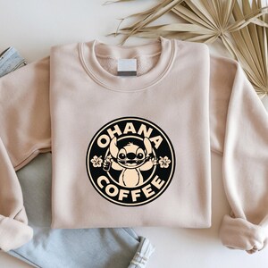 May include: A beige sweatshirt with a circular graphic featuring the text "OHANA COFFEE" and a cartoon character. The graphic is black and tan. The sweatshirt is folded on top of a pair of blue jeans.