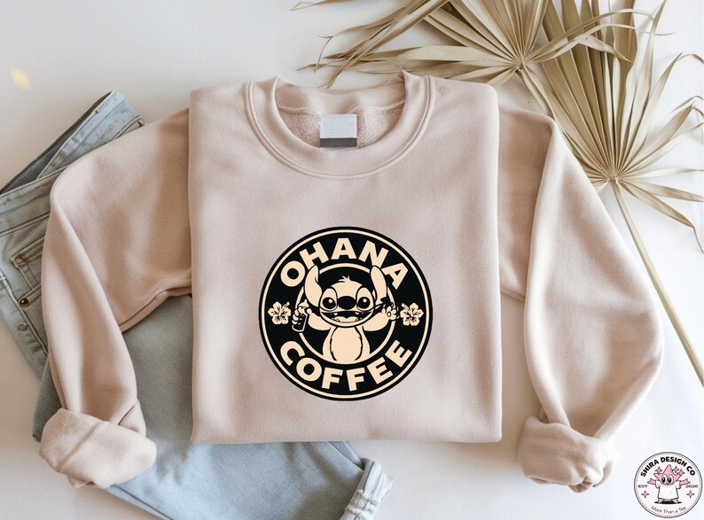 May include: A beige sweatshirt with a circular graphic featuring the text "OHANA COFFEE" and a cartoon character. The graphic is black and tan. The sweatshirt is folded on top of a pair of blue jeans.