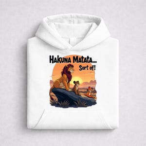 Peut inclure: Sweat à capuche blanc avec un motif coloré de lions sur un rocher au coucher du soleil. Le texte "Hakuna Matata... Sort of!" est au-dessus de l'image. Le motif comprend un lion, une lionne et un lionceau.