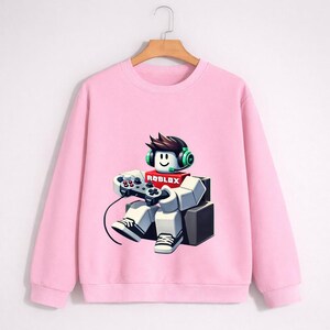 Pu&ograve; includere: Una felpa rosa chiaro con una grafica di un personaggio di Roblox con cuffie e controller. Il personaggio &egrave; seduto e indossa una sciarpa rossa con il logo Roblox. La felpa ha maniche lunghe e girocollo.