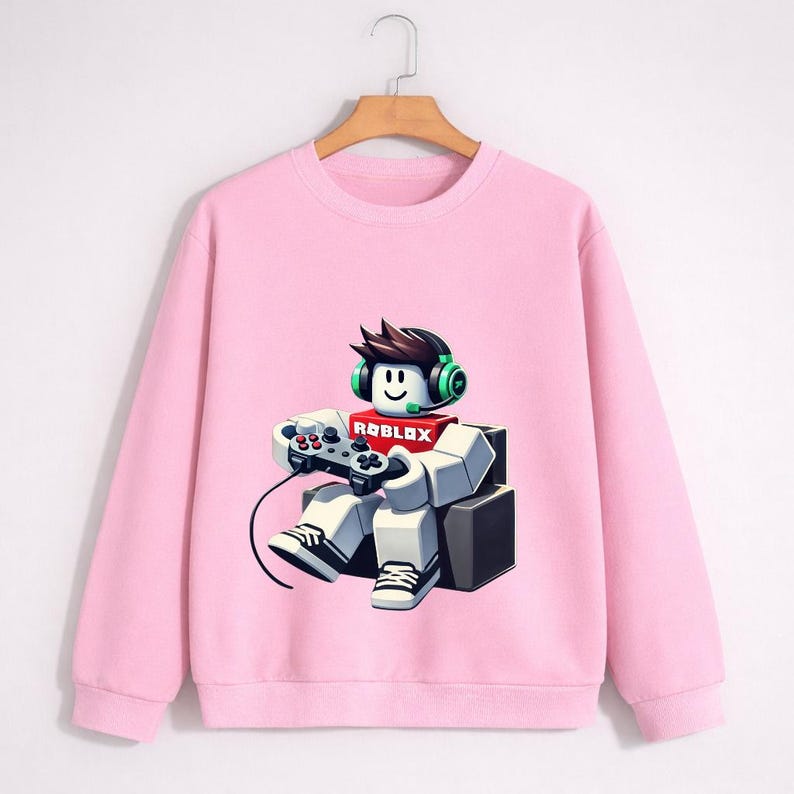 Pu&ograve; includere: Una felpa rosa chiaro con una grafica di un personaggio di Roblox con cuffie e controller. Il personaggio &egrave; seduto e indossa una sciarpa rossa con il logo Roblox. La felpa ha maniche lunghe e girocollo.
