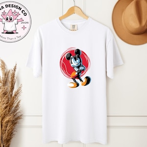 May include: White t-shirt with a cartoon mouse character illustration. The mouse has black ears, a blue body, and orange shorts, set against a red circular background. The shirt is hanging on a wooden hanger.