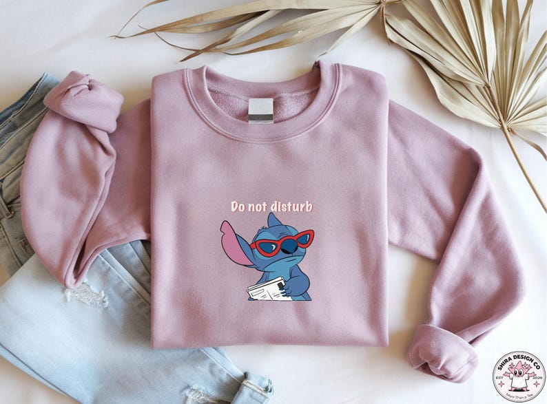 Peut inclure: Un sweat-shirt mauve avec un personnage de dessin anim&eacute; Stitch portant des lunettes de soleil rouges et tenant un journal. Le texte "Do not disturb" est au-dessus du personnage. Le sweat-shirt est pli&eacute; &agrave; c&ocirc;t&eacute; d'un jean bleu.