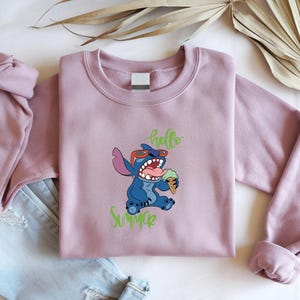May include: A mauve sweatshirt with a blue cartoon character wearing sunglasses and holding ice cream. The words "hello" and "Summer" are written in green script. The sweatshirt is folded next to a pair of blue jeans.