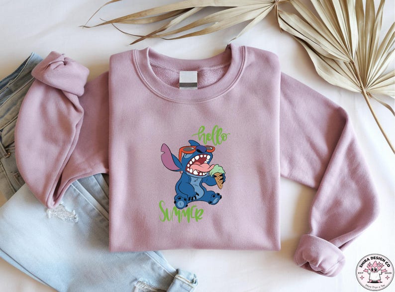 May include: A mauve sweatshirt with a blue cartoon character wearing sunglasses and holding ice cream. The words "hello" and "Summer" are written in green script. The sweatshirt is folded next to a pair of blue jeans.