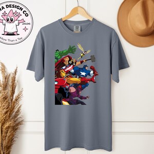 Może przedstawiać: Szara koszulka z kolorową grafiką przedstawiającą postacie z Avengers, w tym Iron Mana, Kapitana Amerykę i Thora. Koszulka wisi na drewnianym wieszaku. Projekt zawiera r&oacute;wnież tekst "Shira Design Co" i "More Than a Tee".