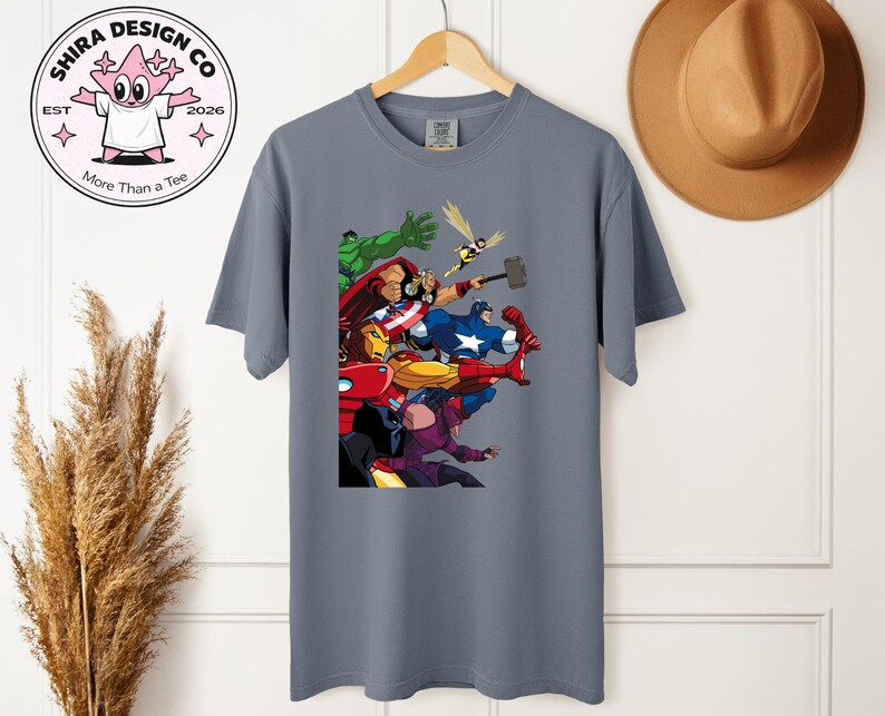 Może przedstawiać: Szara koszulka z kolorową grafiką przedstawiającą postacie z Avengers, w tym Iron Mana, Kapitana Amerykę i Thora. Koszulka wisi na drewnianym wieszaku. Projekt zawiera r&oacute;wnież tekst "Shira Design Co" i "More Than a Tee".