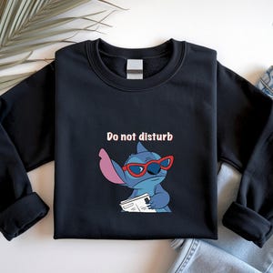 Peut inclure: Sweat-shirt noir avec un personnage de dessin anim&eacute; portant des lunettes rouges et tenant un journal. Le texte "Do not disturb" est au-dessus du personnage. Le sweat-shirt est pr&eacute;sent&eacute; sur une surface blanche avec une feuille de palmier et un jean.