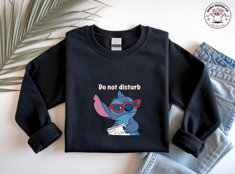 Peut inclure: Sweat-shirt noir avec un personnage de dessin anim&eacute; portant des lunettes rouges et tenant un journal. Le texte "Do not disturb" est au-dessus du personnage. Le sweat-shirt est pr&eacute;sent&eacute; sur une surface blanche avec une feuille de palmier et un jean.