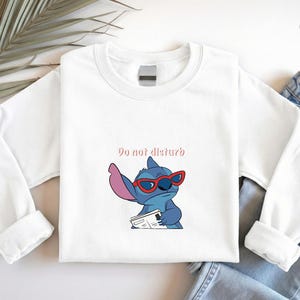 Peut inclure: Sweat-shirt blanc &agrave; manches longues avec une illustration de Stitch portant des lunettes de soleil rouges et tenant un journal. Le texte "Do not disturb" est au-dessus du personnage. Le sweat-shirt est associ&eacute; &agrave; un jean bleu.