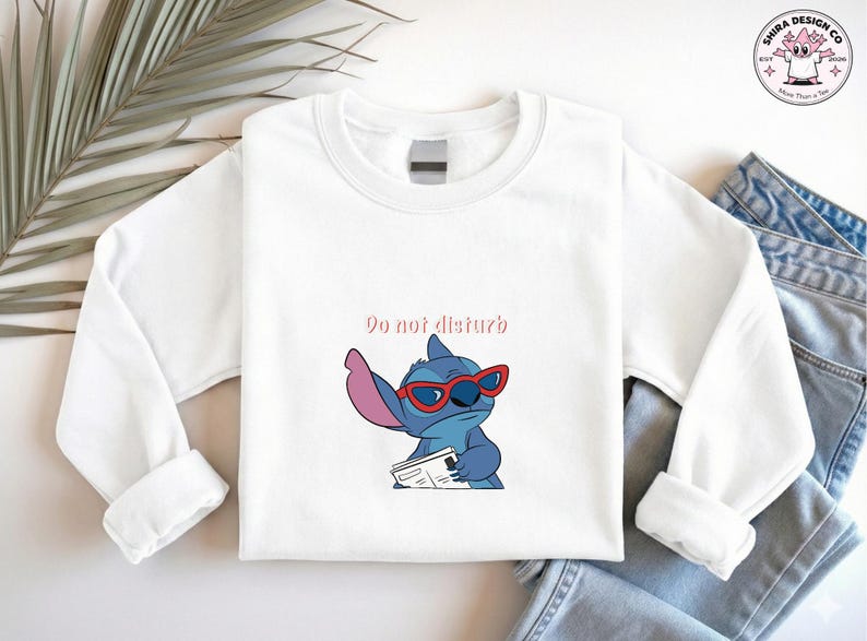 Peut inclure: Sweat-shirt blanc &agrave; manches longues avec une illustration de Stitch portant des lunettes de soleil rouges et tenant un journal. Le texte "Do not disturb" est au-dessus du personnage. Le sweat-shirt est associ&eacute; &agrave; un jean bleu.