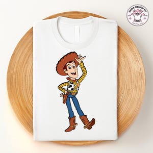 May include: White t-shirt featuring a cartoon illustration of a cowboy character in a brown hat, yellow plaid shirt, blue jeans, and brown boots. The character is smiling and waving. The shirt is folded on a wooden tray.