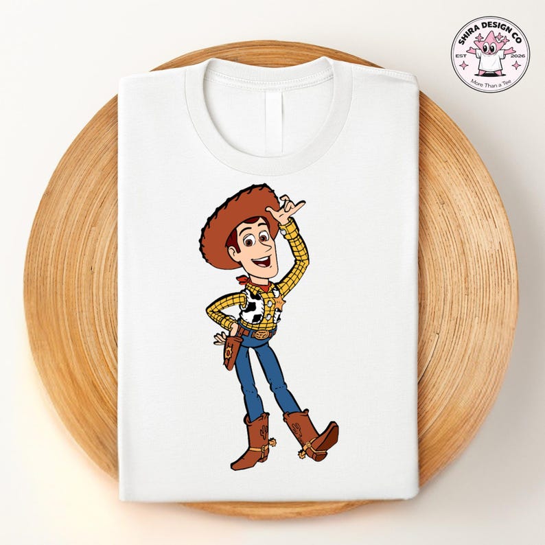 May include: White t-shirt featuring a cartoon illustration of a cowboy character in a brown hat, yellow plaid shirt, blue jeans, and brown boots. The character is smiling and waving. The shirt is folded on a wooden tray.
