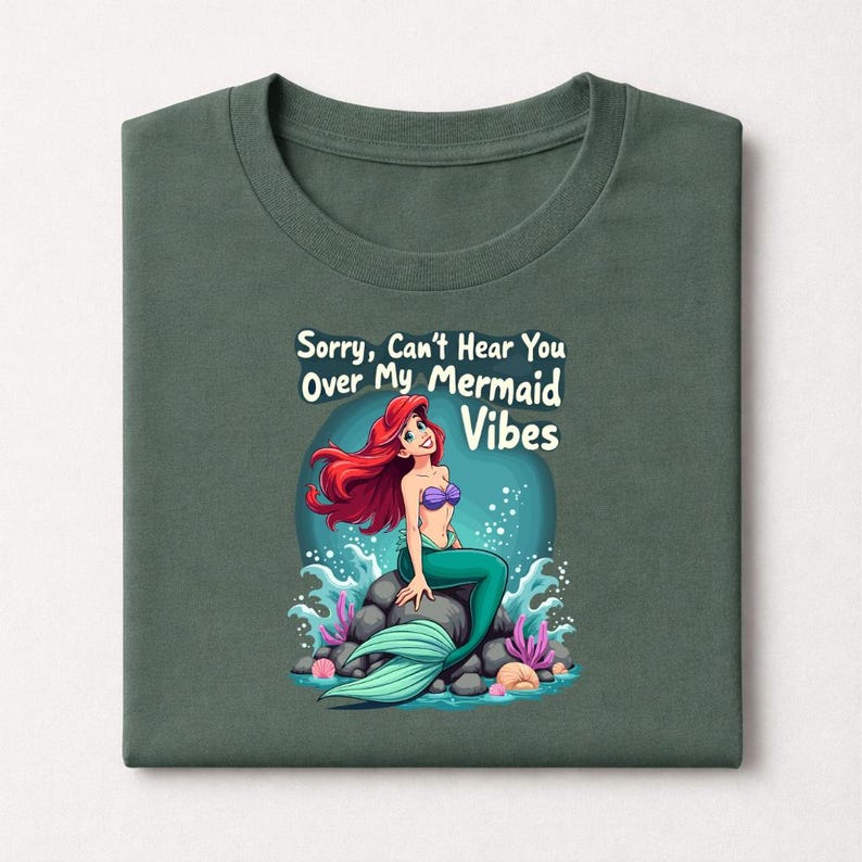 Pu&ograve; includere: T-shirt verde con un design a cartoni animati di una sirena. La sirena ha i capelli rossi, un top viola e una coda verde, seduta su una roccia. Il testo recita "Sorry, Can't Hear You Over My Mermaid Vibes".
