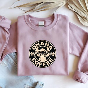 May include: A mauve crewneck sweatshirt featuring a circular graphic with the text "OHANA COFFEE" and a cartoon character. The design is in black and cream. The sweatshirt is folded on top of a pair of blue jeans.