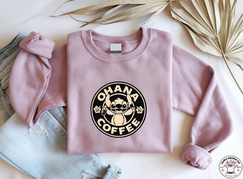 May include: A mauve crewneck sweatshirt featuring a circular graphic with the text "OHANA COFFEE" and a cartoon character. The design is in black and cream. The sweatshirt is folded on top of a pair of blue jeans.