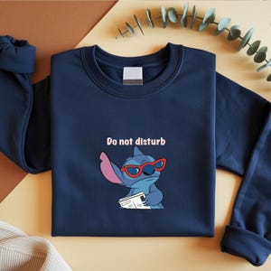 Peut inclure: Sweat-shirt bleu marine avec un personnage de dessin anim&eacute; Stitch portant des lunettes de soleil rouges et tenant un journal. Le texte "Do not disturb" est au-dessus du personnage. Le sweat-shirt a un col rond et des manches longues.