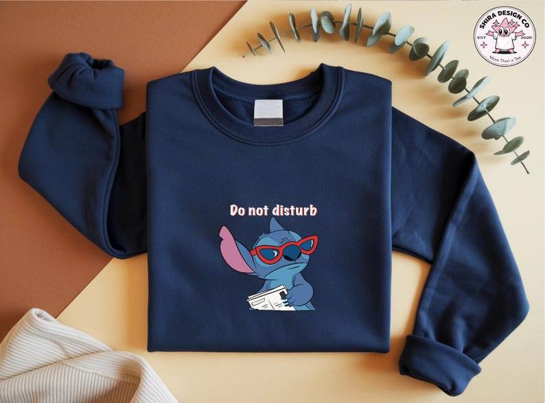 Peut inclure: Sweat-shirt bleu marine avec un personnage de dessin anim&eacute; Stitch portant des lunettes de soleil rouges et tenant un journal. Le texte "Do not disturb" est au-dessus du personnage. Le sweat-shirt a un col rond et des manches longues.