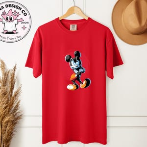May include: A red t-shirt with a cartoon Mickey Mouse illustration. The design is set within a red circle. The shirt is hanging on a wooden hanger. The logo "Shira Design Co" is visible in the upper left corner. A brown hat is visible.