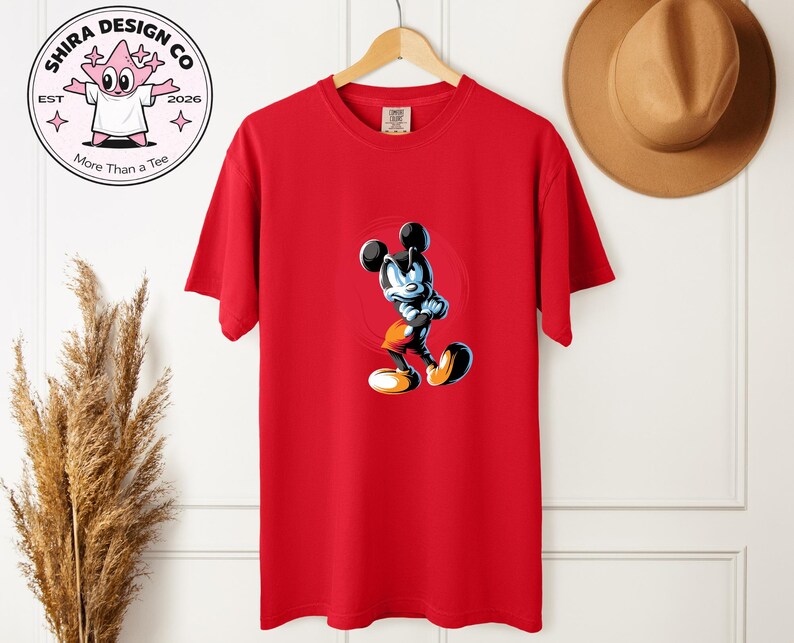 May include: A red t-shirt with a cartoon Mickey Mouse illustration. The design is set within a red circle. The shirt is hanging on a wooden hanger. The logo "Shira Design Co" is visible in the upper left corner. A brown hat is visible.