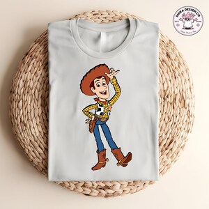 May include: A light gray t-shirt featuring a cartoon illustration of Woody from Toy Story. Woody is depicted in his cowboy attire, including a brown hat, yellow plaid shirt, blue jeans, and brown boots. The shirt has a crew neck.